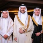 Three Middle Eastern men wearing traditional thobes indoors, smiling and posing.