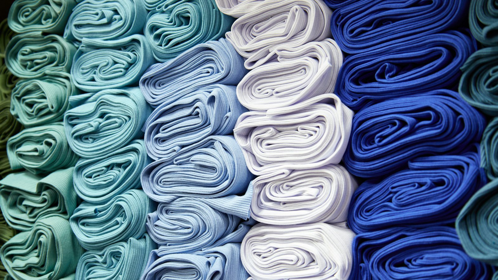 Folded polyester thobe fabric rolls in white, blue and grey on a warehouse shelf